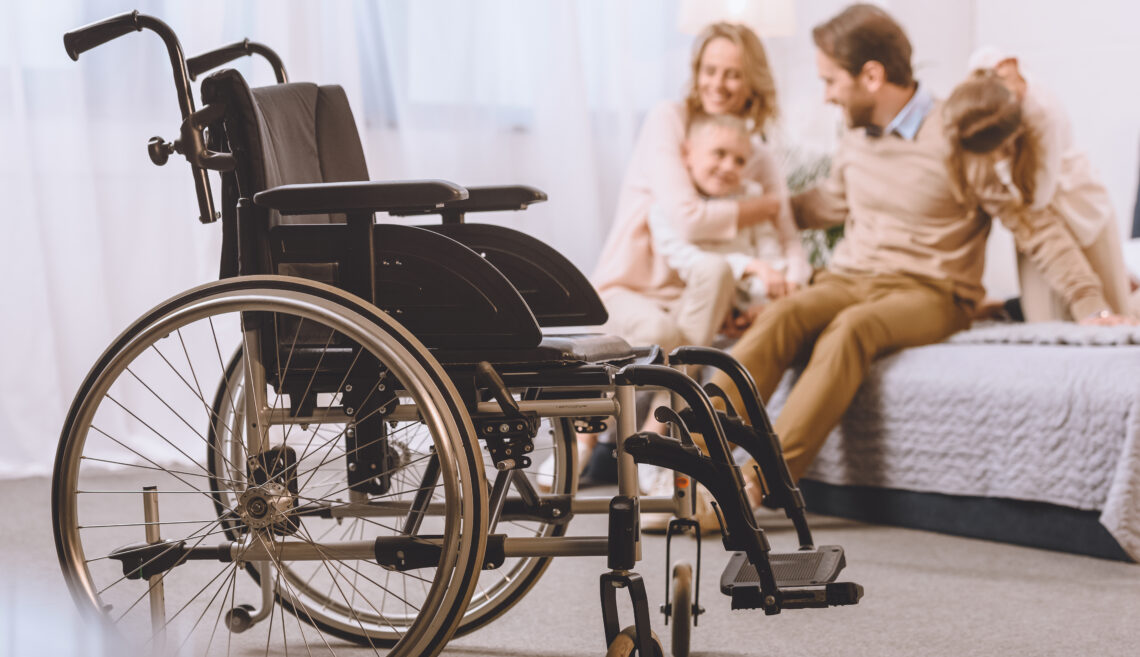 happy father with disability and mother sitting with children on bed with wheelchair on foreground